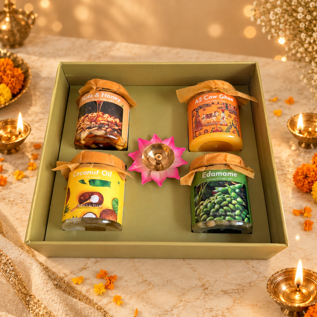 A gift box containing four jars of Ghee & Oils, arranged on a decorated table with marigold flowers and lit diyas. The jars are labeled 'Ghee & Honey', 'AS Cow Curd', 'Coconut Oil', and 'Edamame'.