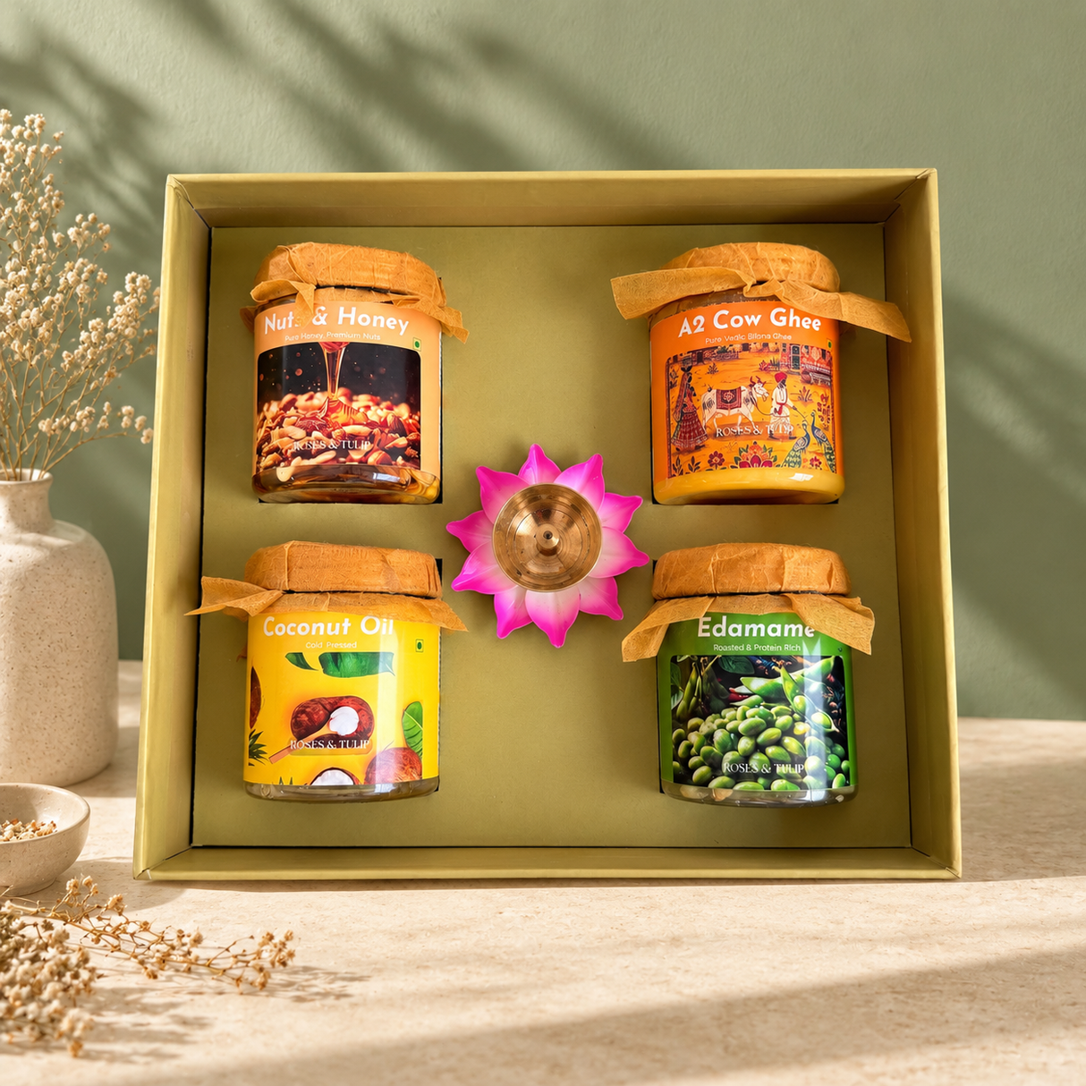 A gift box containing four jars of Ayurvedic and wellness products: Nut & Honey, A2 Cow Ghee, Coconut Oil, and Edamam. The jars are arranged inside a square olive green box with a small pink lotus flower and brass bell in the center.