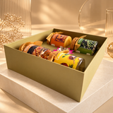 A square olive green gift box filled with assorted packaged snacks and coffee, styled on a light stone pedestal with warm sunlight and decorative dried flowers in the background.
